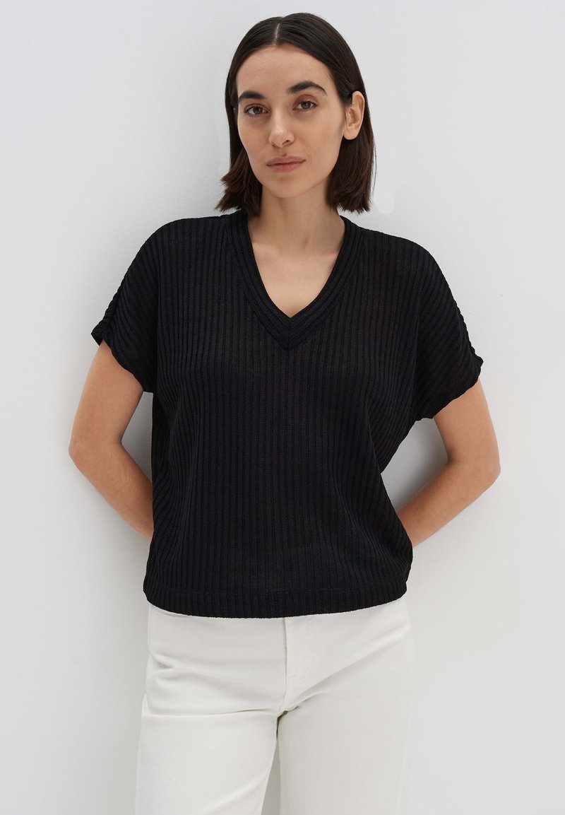 Schwarzes, geripptes kurzärmliges Oberteil mit einem V-Ausschnitt. Der Stoff hat eine strukturierte Oberfläche. Getragen mit hellen Hosen, die Hände auf den Hüften.