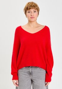 Pull en maille rouge, surdimensionné, avec un décolleté en V profond et des poignets côtelés, porté avec un jean gris. Texture douce, présentant une coupe décontractée.