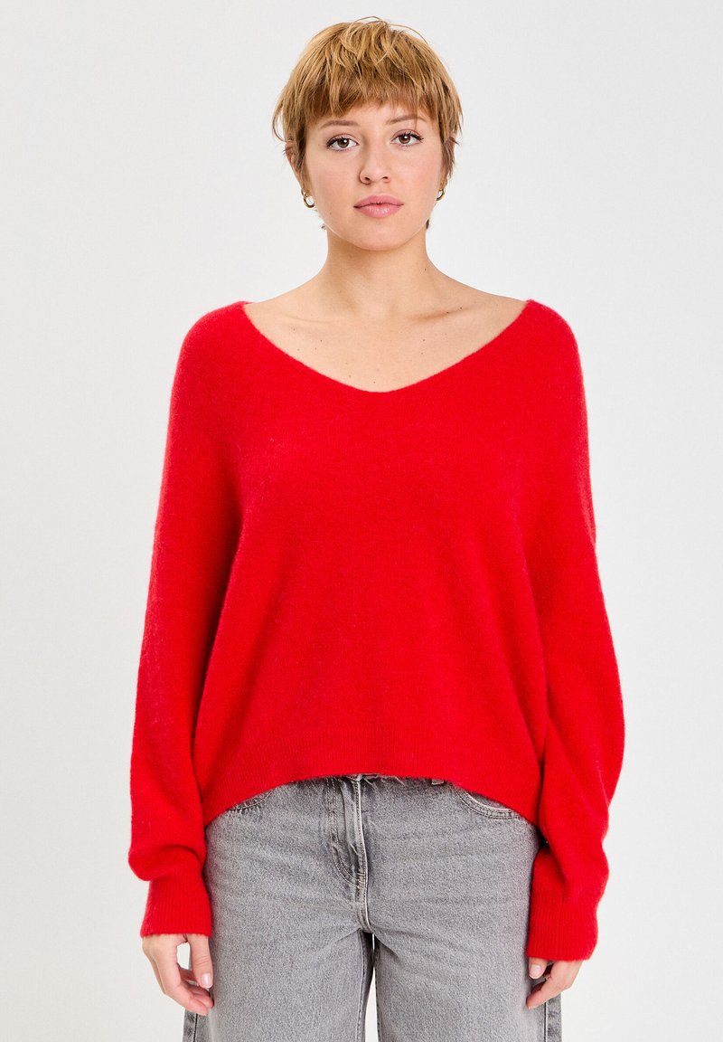 Pull en maille rouge, surdimensionné, avec un décolleté en V profond et des poignets côtelés, porté avec un jean gris. Texture douce, présentant une coupe décontractée.