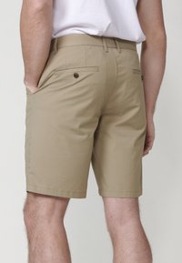 Pantaloni scurți din bumbac bej cu o textură netedă. Au două buzunare la spate, unul cu închidere cu nasture, și buzunare laterale pentru mâini.
