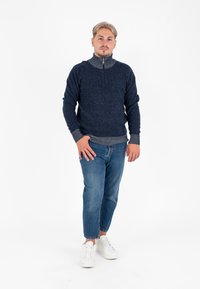 Pullover blu navy con colletto alto e zip, in tessuto a maglia strutturata, abbinato a jeans azzurri e sneakers bianche.