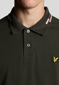 Mörkgrön pikétröja med en strukturerad tyg, som har en randig krage i flera färger och en liten gul örnlogotyp på bröstet.