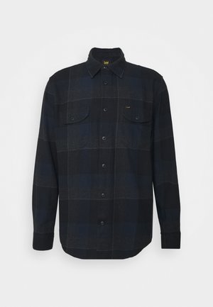 Chemise à carreaux noire et bleu foncé à manches longues, boutonnée, avec col et deux poches poitrine, présentée sur un fond clair uni.
