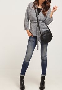 Žena v šedém kardiganu s páskem, vybledlých skinny džínách, černých kotníkových botách a kulaté černé kabelce přes rameno.