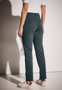 Pantalones verde oscuro de cintura alta, ajustados, con bolsillos traseros y una textura suave, combinados con zapatillas blancas con un acento burdeos.