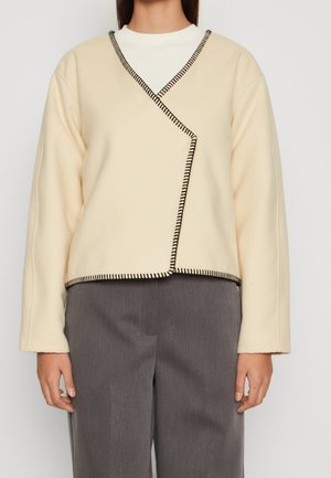 Kermakeltainen, kietaisumallinen takki mustilla yksityiskohdilla, pitkät hihat ja pehmeä rakenne, yhdistetty harmaisiin housuihin.