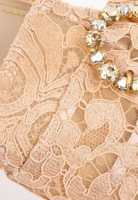 Tissu en dentelle beige avec des motifs floraux complexes et une bague en or ornée de gros strass clairs dans le coin supérieur droit.