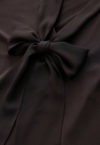 Tissu noir à texture lisse avec un détail de nœud attaché au centre. Le matériau semble léger et fluide.