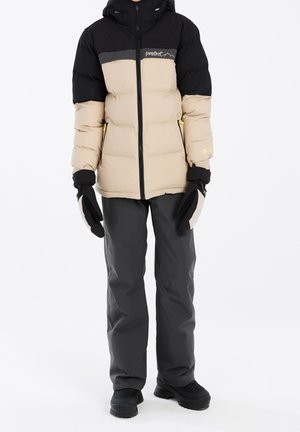 Doudoune avec une partie supérieure noire et une partie inférieure beige, dotée d'une fermeture éclair avant, d'une capuche et d'accents jaunes. Associée à un pantalon gris et des bottes noires.