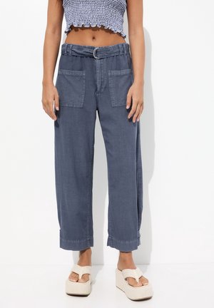 Mujer con pantalones azules de pierna ancha y talle alto, con grandes bolsillos delanteros, cinturón a juego, top corto con volantes y sandalias blancas de plataforma.