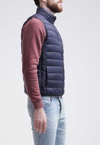 Námořnický modrý prošívaný vestek s vysokým límcem, prošívaným designem a bočními kapsami, nošený přes vínově červenou dlouhým rukávem košili a modré džíny.