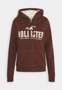 Brun zip-up hoodie med framfickor, vit "Hollister Southern California" logotyp, dragsko huva och mjuk vit foder inuti huvan.