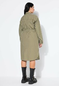 Olivengrün gestreiftes Hemdkleid mit langen Ärmeln, tailliertem Bund, Seitenschlitzen und kontrastierenden weißen Streifen. Kombiniert mit knielangen schwarzen Stiefeln.