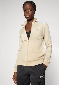 Beige zip-up jacket with high collar, white shoulder accents, and Puma logo. Worn with dark pants, showing smooth, soft fabric texture.