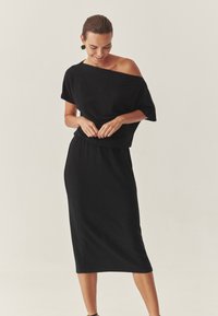 Rochie tricotată neagră cu un design off-the-shoulder, mâneci scurte și talie ajustată, cu o textură netedă și tiv de lungime medie.