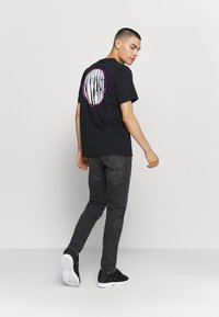 Jovem a caminhar, usando ténis pretos, jeans escuros e uma t-shirt preta com um grande logótipo circular branco e rosa nas costas.
