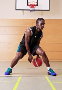 Zwarte mouwloze sporttop met witte accenten, zwarte shorts en multicolored basketbalschoenen. Dribbelt een rode basketbal op een gymvloer.