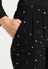 Zwarte broek met een gladde textuur en kleine witte driehoekige patronen. Heeft een elastische tailleband en een zijzak.