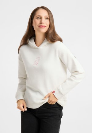Smiling woman with long brown hair wearing a white hoodie with a small pink lightning bolt design and black pants, hands in pockets.