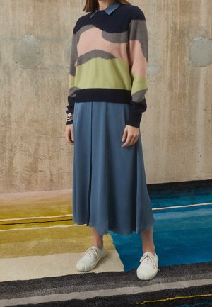 Person mit einem mehrfarbigen Pullover, blauem Midirock, weißen Turnschuhen und Perlenarmbändern, die auf einem mehrfarbigen Teppich vor einer Betonwand steht.