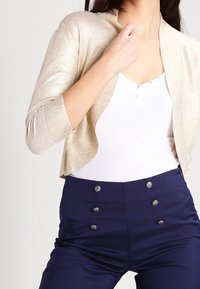 Gouden glinsterende cropped cardigan over een wit aansluitend topje, gecombineerd met marineblauwe hoge taillebroek met zilveren knoopaccenten.