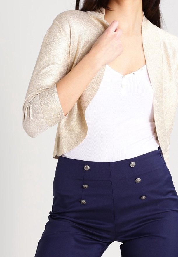 Gouden glinsterende cropped cardigan over een wit aansluitend topje, gecombineerd met marineblauwe hoge taillebroek met zilveren knoopaccenten.