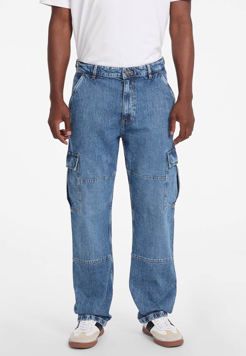 Calças de ganga cargo de azul claro com pernas largas, dois bolsos laterais e detalhes de costura únicos. Usadas com uma t-shirt branca e sapatilhas.