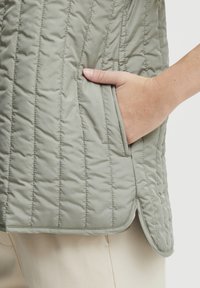 Gilet matelassé vert clair avec des coutures verticales, doté d'une poche latérale. Le matériau semble lisse avec un léger éclat et un ourlet arrondi.