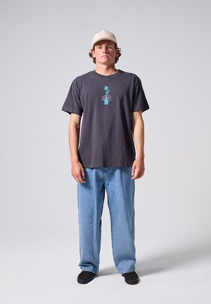 Tânăr în picioare, purtând o șapcă bej, tricou închis cu imprimeu cu floare albastră, blugi deschis la culoare și pantofi negri, pe un fundal simplu.