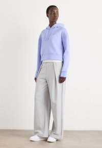 Haut de survêtement court en lavande clair avec capuche à cordon et logo, associé à un pantalon de jogging gris à jambes larges et des baskets blanches. Textures lisses.