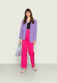 Blazer lavanda sobre uma blusa preta, combinado com calças rosa-choque de cintura alta. Os acessórios incluem uma bolsa lilás clara e sandálias combinando.