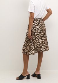 Osoba ubrana w spódnicę midi w leopardzi wzór, białą koszulkę i czarne mule na słupku z odkrytymi palcami, stojąca na tle jednolitej białej ściany.