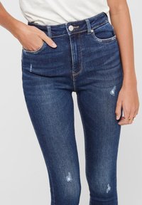 Jeans skinny de culoare albastru închis, cu talie înaltă, cu ușoare pete de decolorare și deteriorare, o mână în buzunar, purtate cu un top alb.
