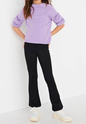 Personne portant un pull violet clair, un pantalon noir évasé et des baskets à plateforme blanches, debout les mains sur les hanches sur un fond blanc.