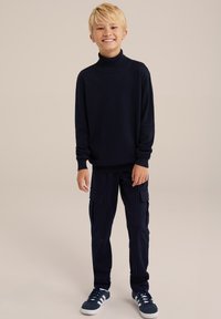 Mornarsko modra turtleneck pulover z dolgimi rokavi, kombiniran z temnim hlačami cargo z navpičnimi črtami, ki imajo stranske žepe, in temnimi športnimi čevlji.