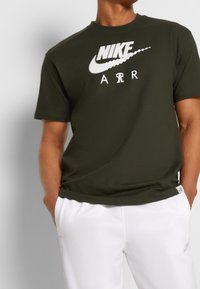 Krátké olivově zelené tričko Nike s krátkým rukávem, zdobené velkým bílým logem a nápisem "AIR", vyrobené z bavlny, s kulatým výstřihem a volnějším střihem.