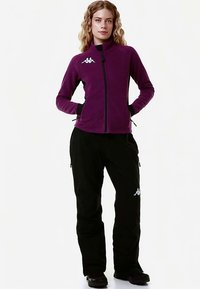 Giacca in pile viola con chiusura a zip e collo alto, abbinata a pantaloni neri con cerniere laterali e logo bianco sulla coscia.