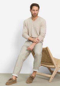 Maglione beige con scollo a V, pantaloni grigio chiaro e scarpe in camoscio marrone con suola in espadrillas. Modello seduto su una sedia di legno intrecciato.