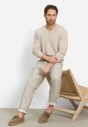 Beige trui met V-hals, lichtgrijze broek en bruine suède schoenen met espadrille-zolen. Model zittend op een gevlochten houten stoel.