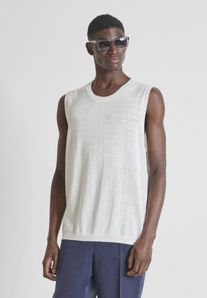 Uomo con top bianco in maglia senza maniche, occhiali da sole scuri e pantaloni blu navy, in piedi davanti a uno sfondo grigio chiaro uniforme.