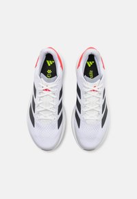 Baskets de sport blanches avec des rayures noires, un dessus en maille pour la respirabilité, et des accents rouge vif sur la languette et le talon.