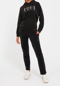 Sudadera negra con texto gráfico frontal y cordón, combinada con pantalones suaves negros a juego. Zapatillas blancas completan el conjunto.