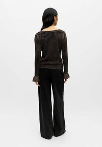 Kvinde med langt sort hår iført en brun mønstret langærmet top og bredbenede sorte bukser, der står med ryggen til mod en hvid baggrund.