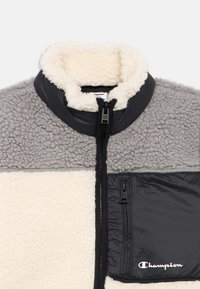 Pilkos ir smėlio spalvos vilnonis džemperis su juodu užtrauktuku krūtinėje ir aukštu apykakle, ant kišenės esantis „Champion“ logotipas.