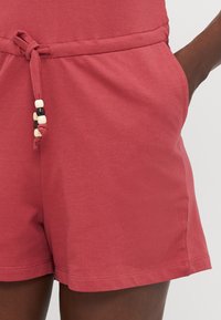 Short rouge décontracté avec un cordon orné de perles noires et blanches, porté par une personne avec une main dans la poche.