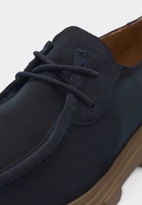 Σκούρο μπλε δερμάτινο παπούτσι με κορδόνια, με ραμμένα σχέδια και ελαφριά καφέ λαστιχένια σόλα, εμφανιζόμενο σε κοντινό πλάνο σε λευκό φόντο.