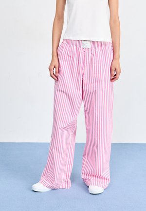 Persona que lleva pantalones sueltos con rayas verticales rosas y blancas, zapatillas blancas y una camiseta sin mangas blanca, de pie sobre un suelo azul.