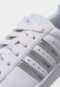 Λευκό αθλητικό παπούτσι Adidas Superstar με ασημί ρίγες, λευκά κορδόνια και ανάγλυφο λογότυπο Adidas στη γλώσσα.