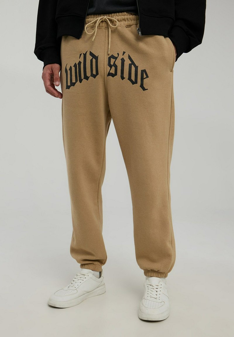 Pantaloni sportivi in cotone beige con "wild side" stampato in nero, dotati di un elastico in vita, cordino e polsini a costine. Indossati con sneakers bianche.