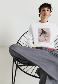 Felpa bianca con una stampa grafica tonalità rosa, abbinata a pantaloni larghi grigi. Il modello è seduto su una sedia nera a rete, con le gambe incrociate.